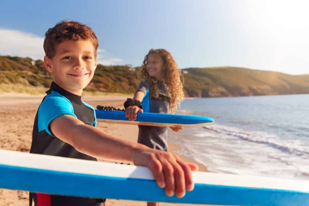 Kid on the beach ready to go into the water with their Bodyboards, article which explains why bodyboarding is a good exercise and if it is also good for kids