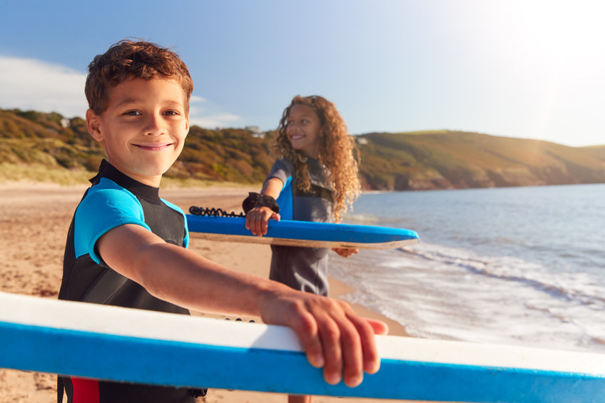 Portrait Of Children Wearing Wetsuits Carrying Bodyboards On Summer Beach Vacation Having Fun By Sea