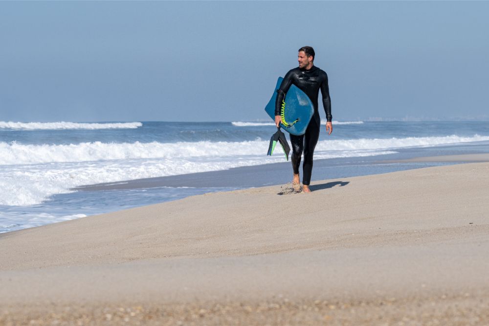 Man walking out the sea with a full body black wetsuit on and holding a bodyboard.