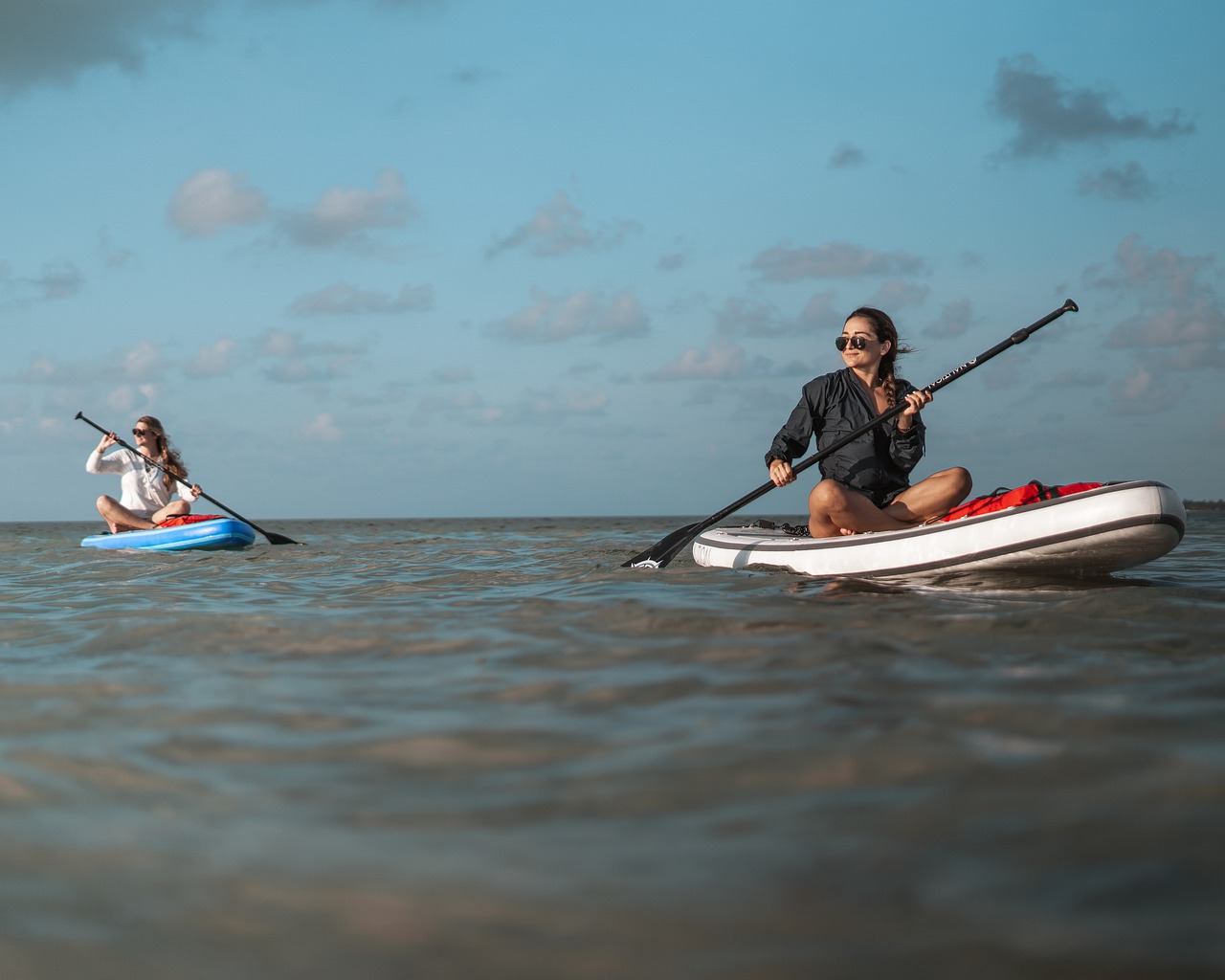 Two women in the ocean sitting on paddleboard whilst looking towards sun