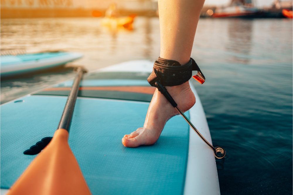 Nahaufnahme eine Frau, welche auf einem blau-gelben Stand Up Paddle oder SUP Board steht und eine SUP Leash trägt Wie man eine SUP Leash am SUP Board anbringt, wie man die richtige Länge einer SUP Leash bestimmt, Arten von SUP Leashes