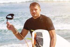 Mann am Strand mit seinem Bodyboard der auf die Bodyboard Leine zeigt