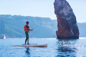 Ein Mann fischt auf einer Angelrute auf einem Stand Up Paddleboard, SUP Angeln, Angeln auf einem SUP, Mann der auf einem Stand-Up-Paddle-Board fischt