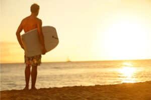 Bodyboarder am Strand im Sonnenuntergang der ein Bodyboard unter dem Arm trägt