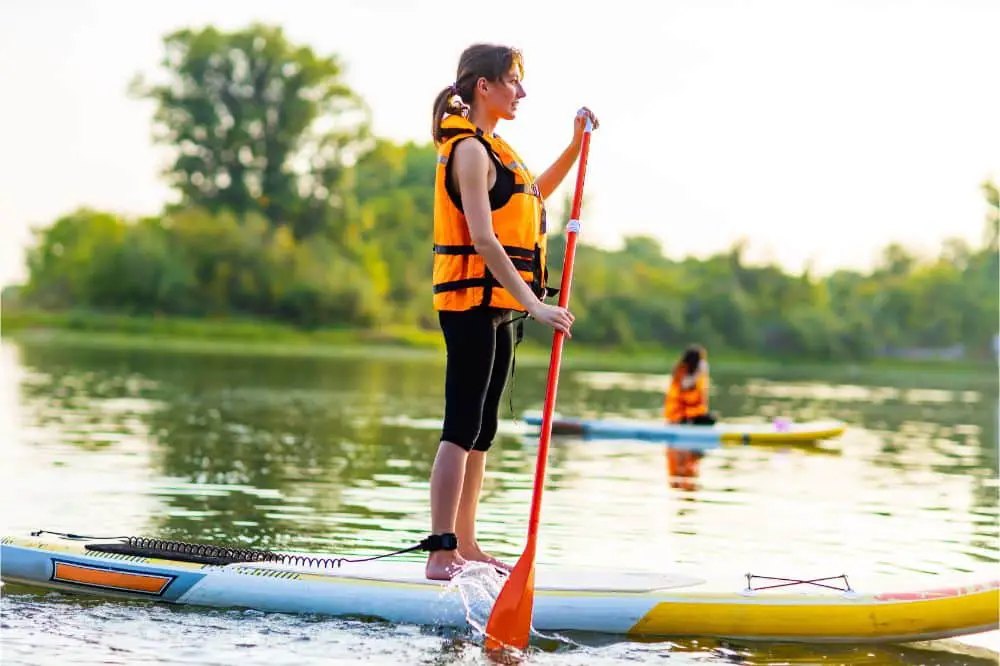 woman wearing life vest paddleboarding, article answering the question Do you need  a life vest on a paddleboard? Woman on a lake on a Stand Up Paddle Board wearing a Life Jacket in an article about the necessity of life jacket and personal floating devices while paddleboarding.