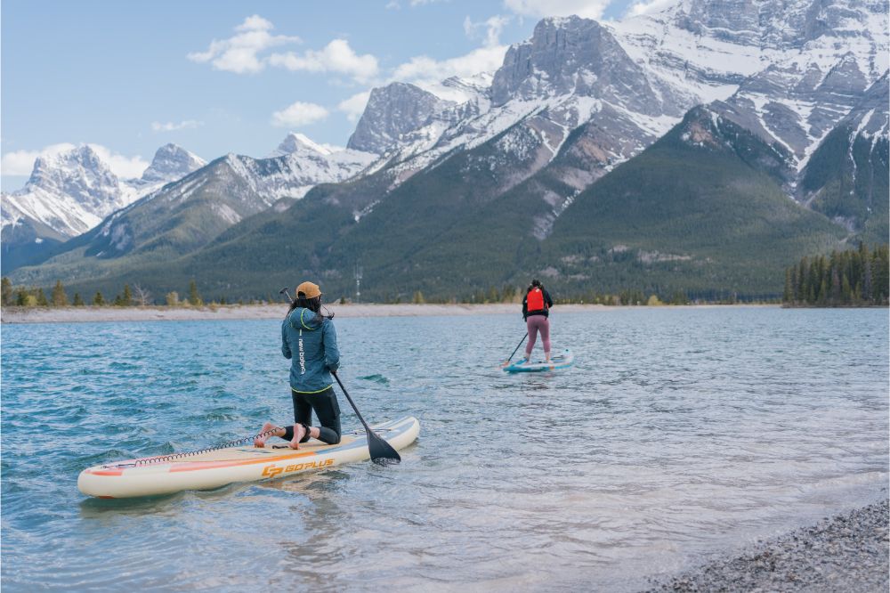 People paddle boarding in shallow water with mountain ranges and trees in the background