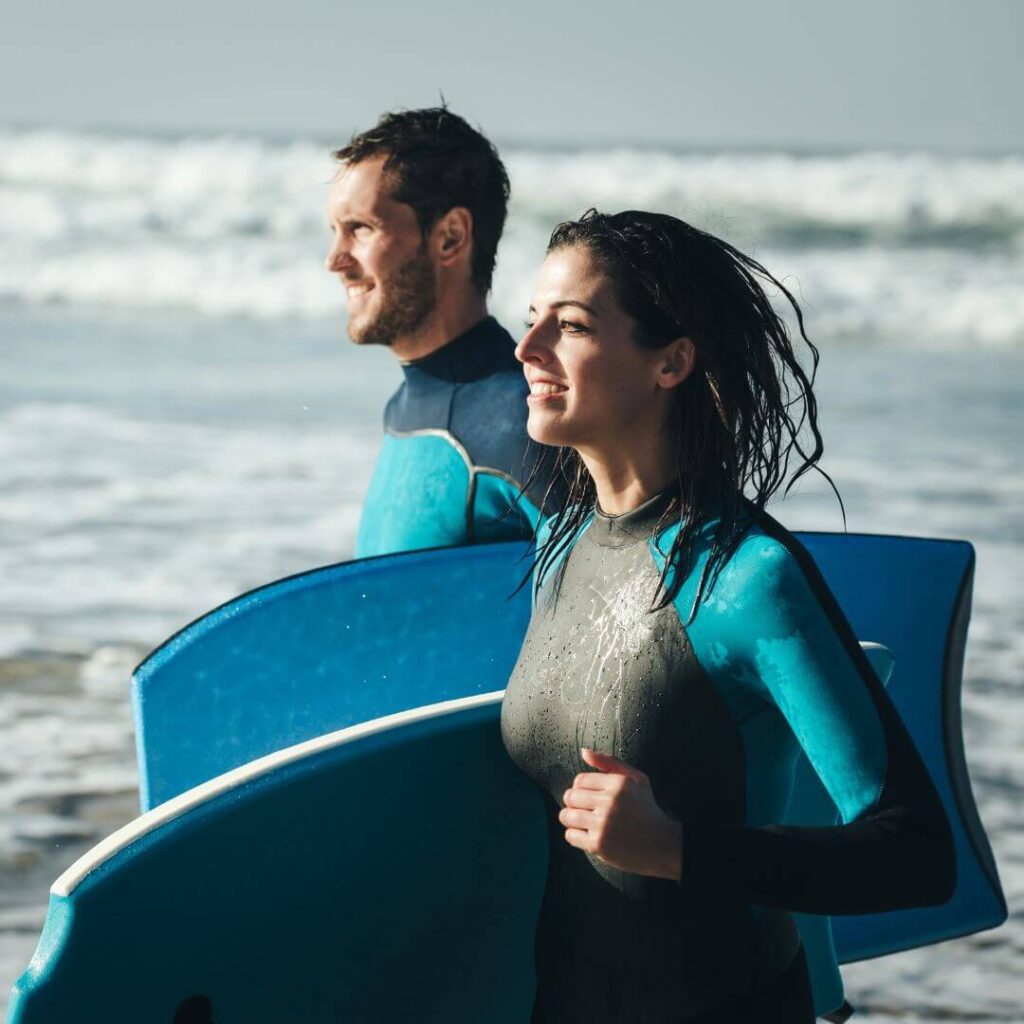 Picture of a man and a woman in wet suits holding body boards running in from the sea with waves in the background