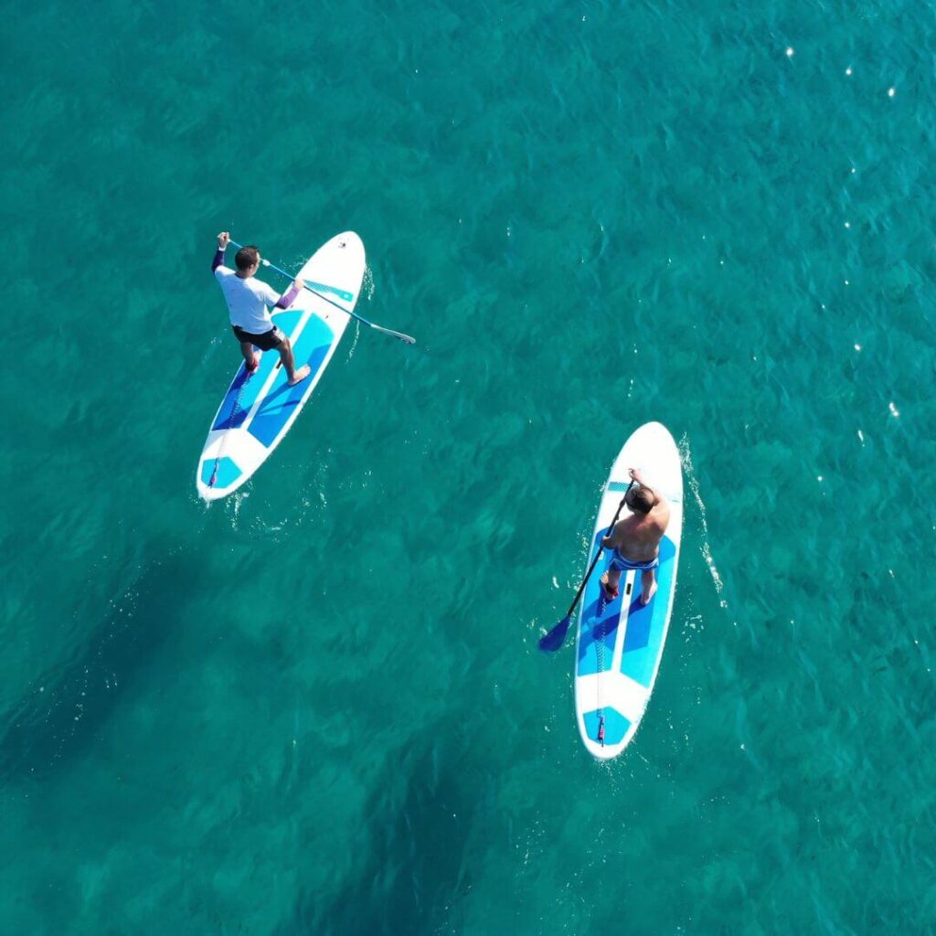 Birds eye view pircture of two guys stood up on surf boards together in the sea holding paddles