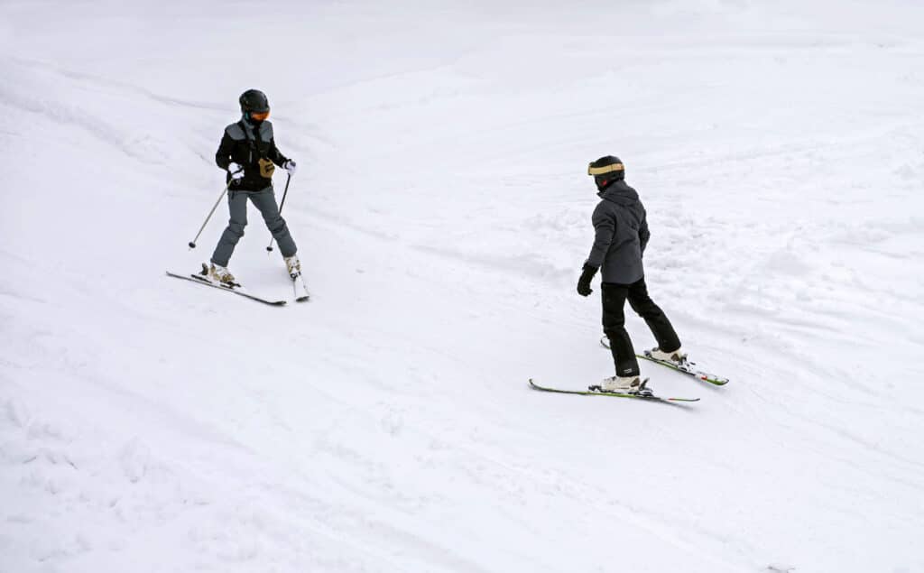 Two people facing each other at the bottom of a ski slope, showcasing 'The Snowplow' skiing technique.
