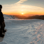 A person at the top of a ski slope, admiring the sun setting behind the mountains.