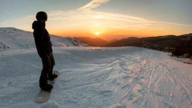 A person at the top of a ski slope, admiring the sun setting behind the mountains.