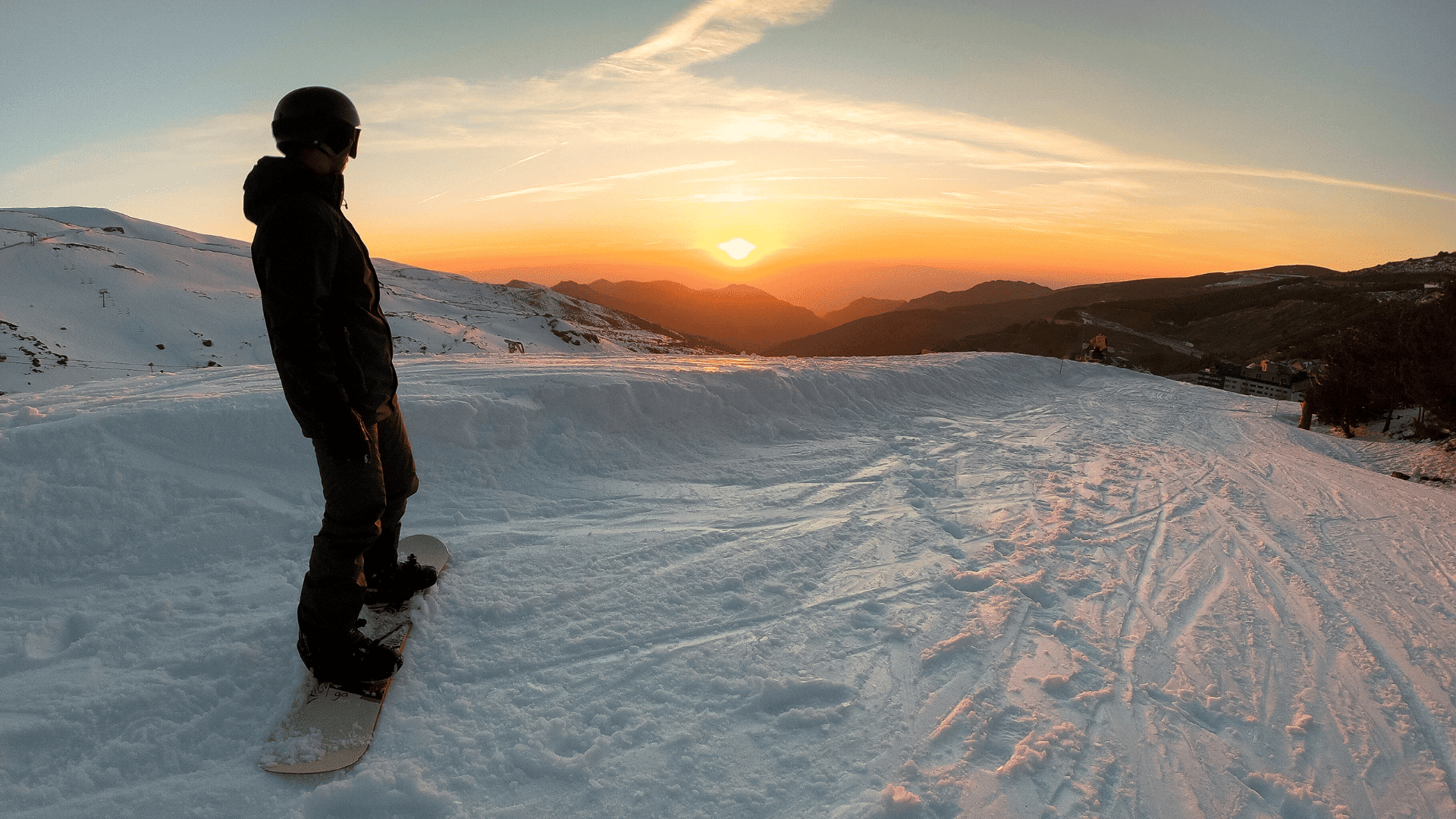 A person at the top of a ski slope, admiring the sun setting behind the mountains.