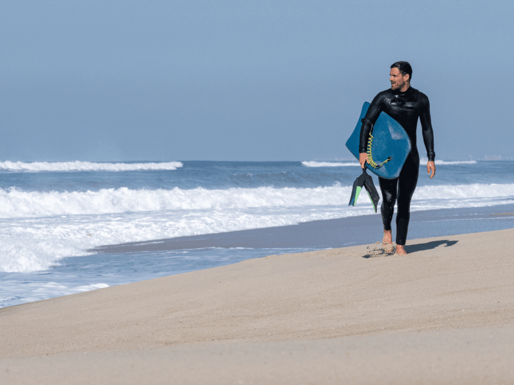 A man in a wetsuit walking across a beach holding a body board
