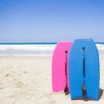 Two body boards, one pink and one blue stood up in the sand on a beach