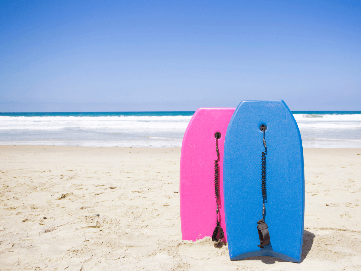 Two body boards, one pink and one blue stood up in the sand on a beach