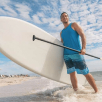 Man standing in a blue tank top and shorts holding a paddle and paddle board standing in the sea on a beach