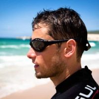 A man on the beach wearing a wetsuit and floating sunglasses looking out into the sea